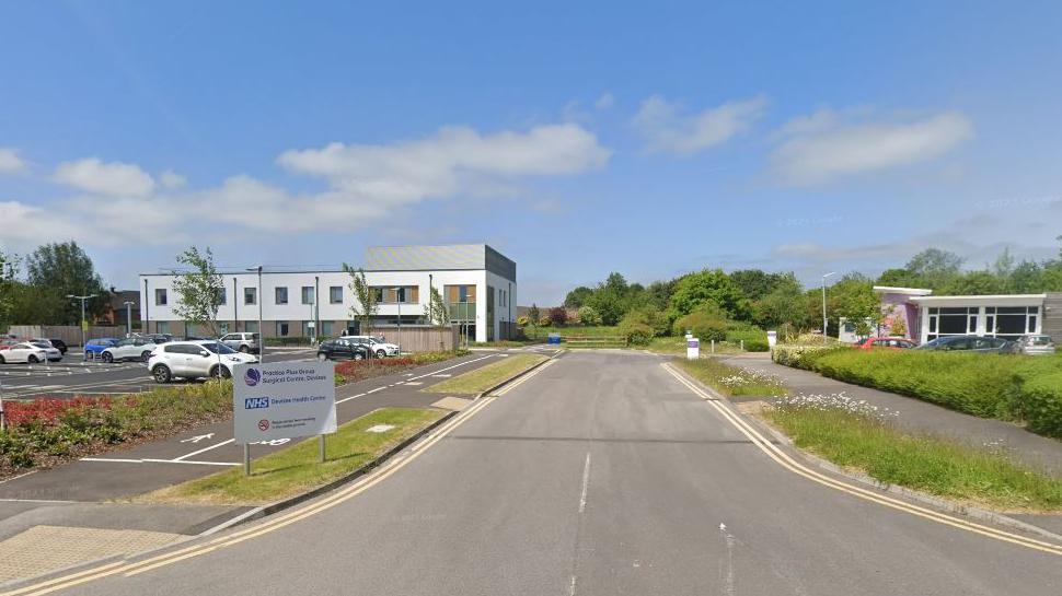 A view of two buildings, including a health centre with car park in front, a road going down the middle with grassy verges under a blue sky.