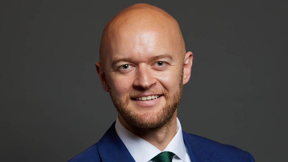 Close-up image of Jonathan Hinder, wearing a blue suit and green tie, and smiling in front of a dark grey background.