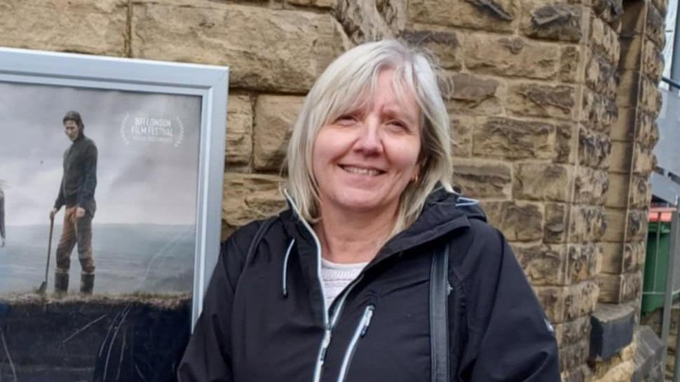 Dawn Lennie, standing in front of a brown wall. She has long white hair and is wearing a black rain jacked with white zips. A film poster is attached to the wall behind her. 