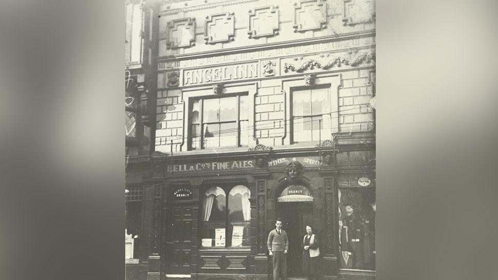 An undated black and white image of the exterior of the Angel Inn in Stockport with a man and a woman standing at the entrance.