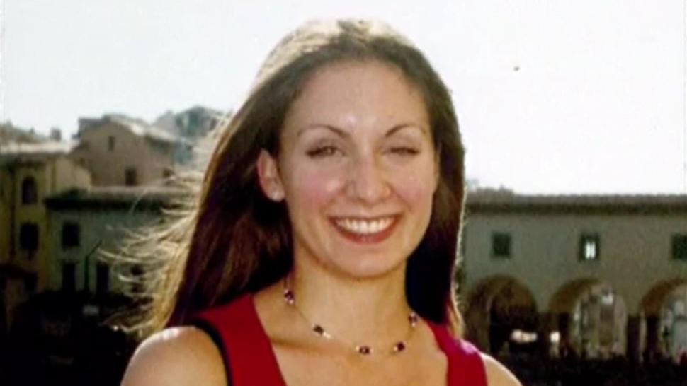 A woman in a red top with long brown hair smiles for the camera whilst on holiday by the sea.