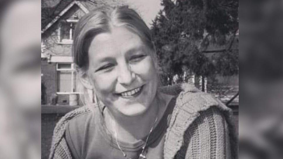 Dawn Sturgess in a black and white photograph. She has blonde hair, is smiling at the camera and squinting into the sunlight. She has a thick cardigan on and a T-shirt. 