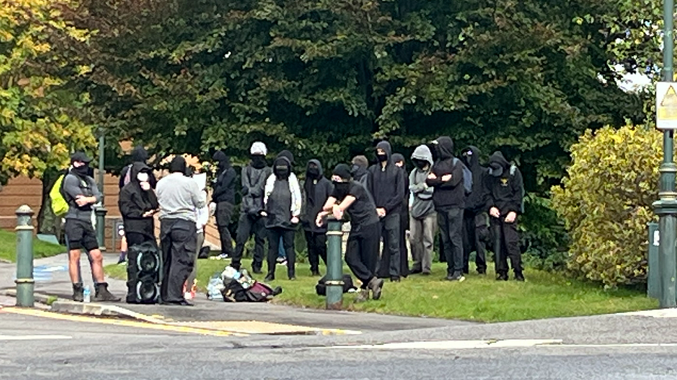 About 20 members of the Antifa group, standing on a grassed area, wearing hoods to cover their heads and many wearing facemasks. To the right of them, there is a sign for the Bournemouth Civic Centre and then, to the right of that, two police officers are either sat or stood looking in another direction