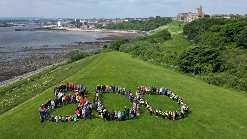 Aerial shot of a view of a river at low tide with buildings, including two white former lighthouses in the distant background, and closer in a large block of flats at the top of a steep bank with grass and trees. In the foreground, there is a flat grassed area with dozens of standing people arranged in the shape of the figure 800.
