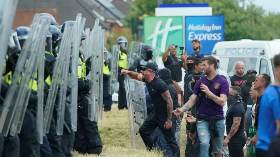 Police line up with riot shields as a group of men stand off with them outside the Holiday Inn Express in Rotherham last August 