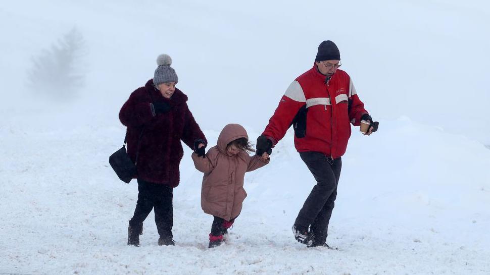 A man and woman, wearing coats and hats, hold hands with a small girl in a coat with her hood up as they walk across snowy ground in Glenshee in Scotland