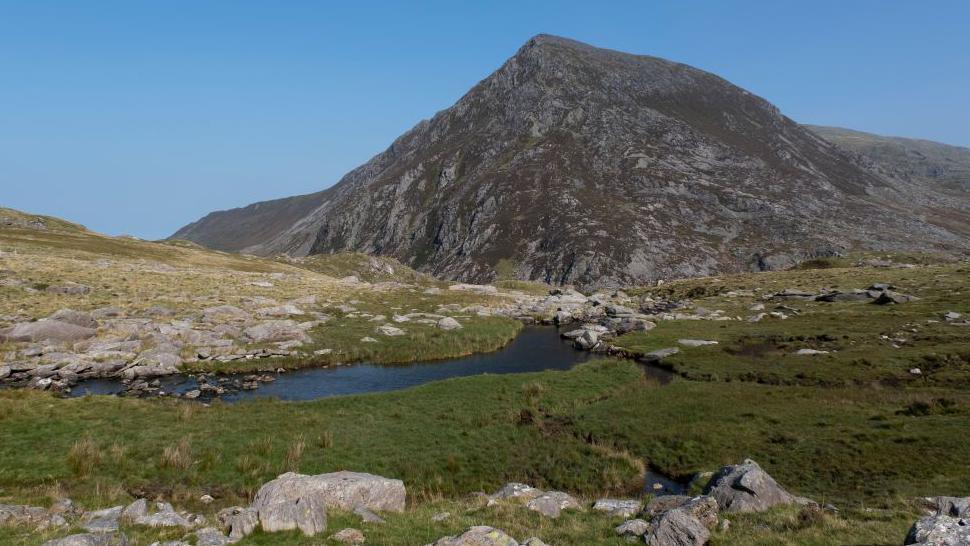 Landscape view looking towards the summit of Pen yr Ole Wen. There's a blue sky behind, with a winding stream in front of the mountain and rocks scattered on the grass around it. 
