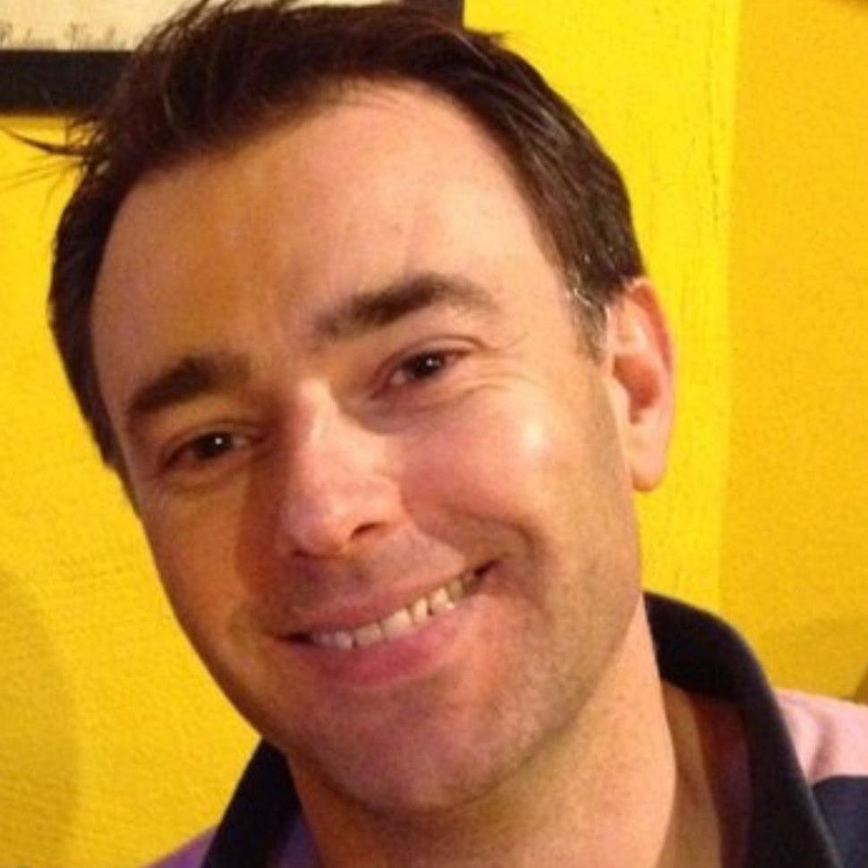A smiling man with short brown hair in a bright yellow room. He is wearing a pink and navy striped polo shirt.