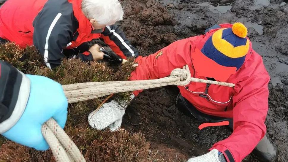 The man, in a red coat and blue and yellow striped bobble hat, waist deep in mud with a rope around the top of his chest, which is held by someone with a blue glove, being assisted by a member of the rescue team with white hair who is bending over to secure a rope. 