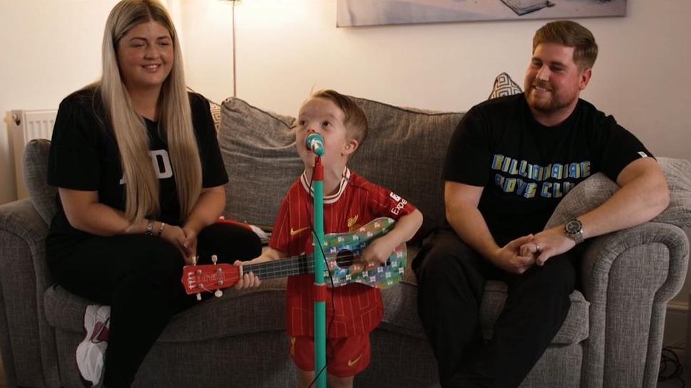 Isaac sings into a toy microphone stand while strumming his colourful striped toy guitar. His mother and father watch while sitting on a sofa behind him.