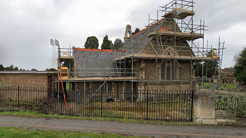 A former chapel in a cemetery. It looks like a small church. It has scaffolding on the outside. 