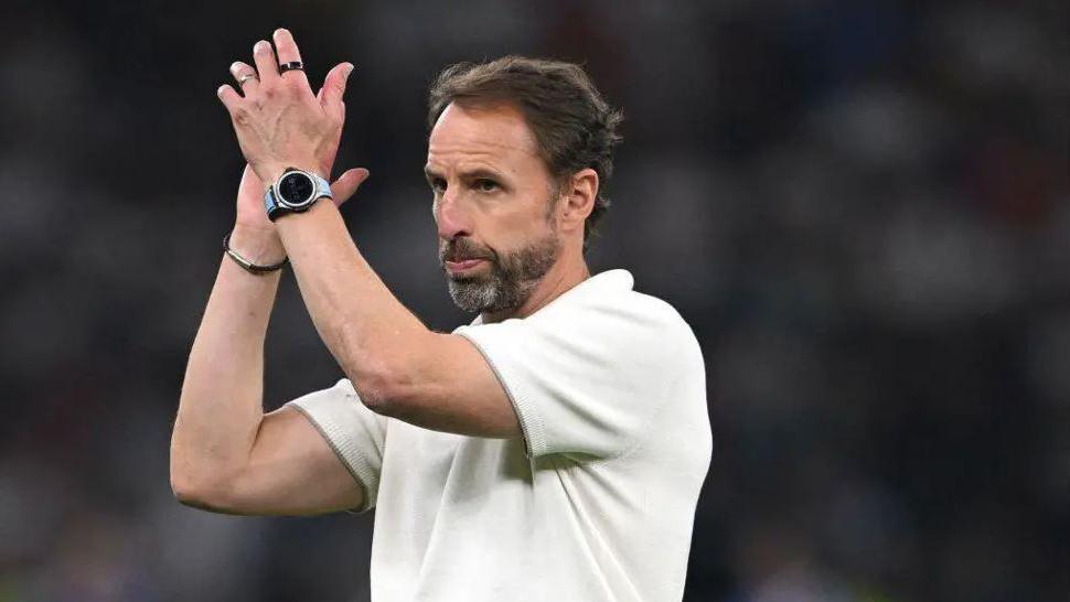 Gareth Southgate claps after a football game. he is wearing a white t-shirt and wears a watch and bracelet. he has brown hair and a greying beard