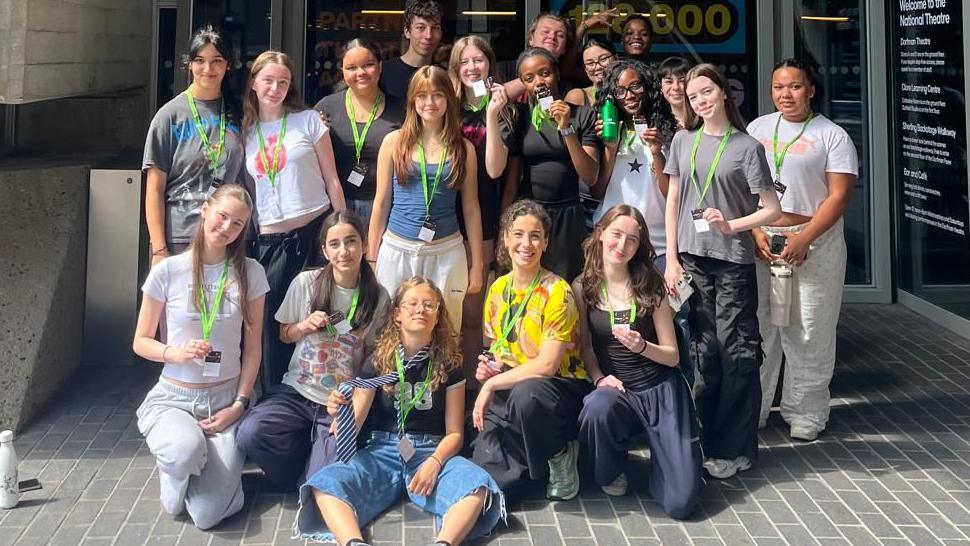 A group of teenagers standing outside the National Theatre in London. The teenagers are standing in rows, with those at the front kneeling or sitting down. They are wearing casual clothes.