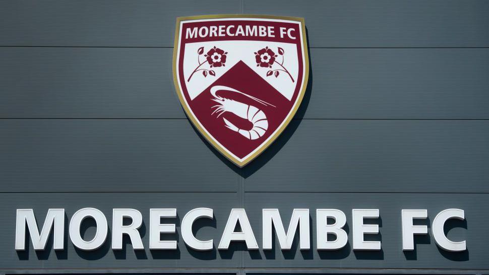 The Morecambe Football Club crest above the name 'Morecambe FC', which is written in capital letters, as seen on the front of their home ground, the Mazuma Mobile Stadium