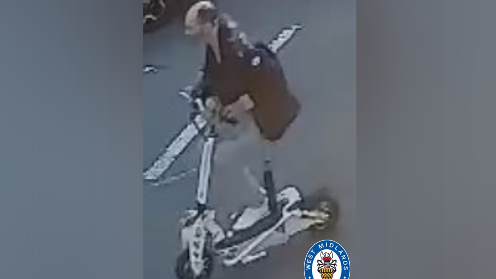 A bold man wearing a black jacket and grey bottoms riding a white e-scooter 