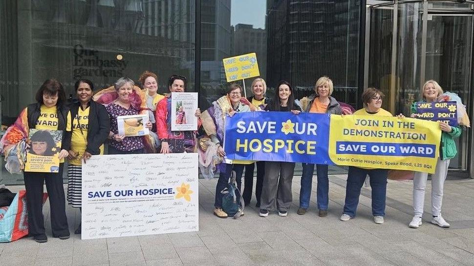 A group of 11 people stood in a line holding blue and yellow banners which say "Save Our Hospice" and "Join in the demonstration save our ward".