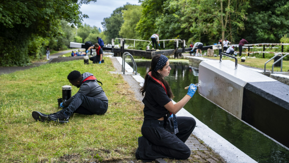 A woman at the side of a canal painting a lock bridge, with a man nearby on some grass fixing a fitting. 