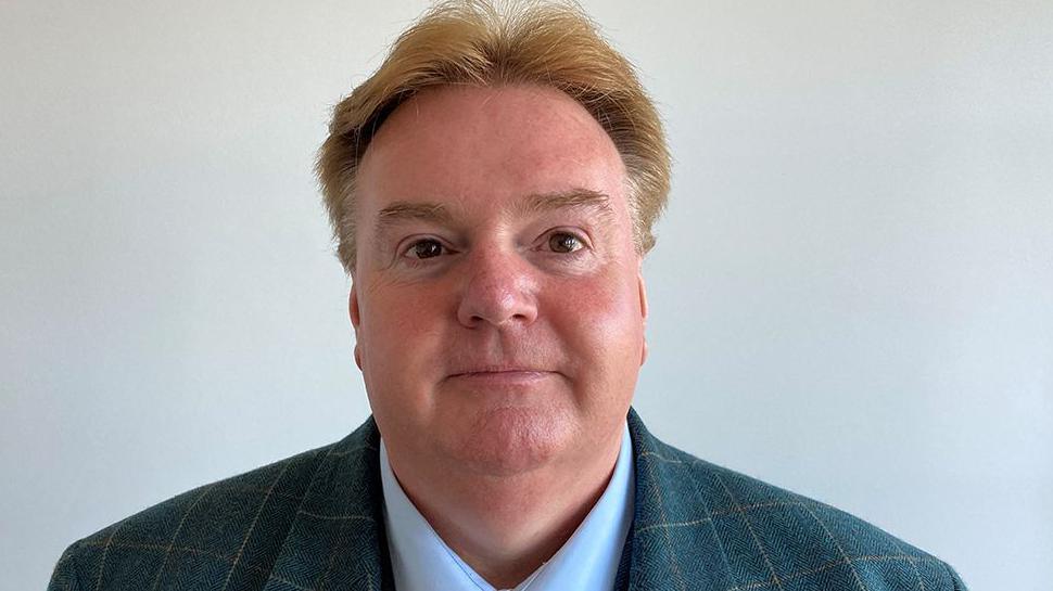 Head and shoulders image of Mark Clifford in front of a plain grey wall. He has sandy hair parted in the middle and is wearing a checked suit and pale blue shirt