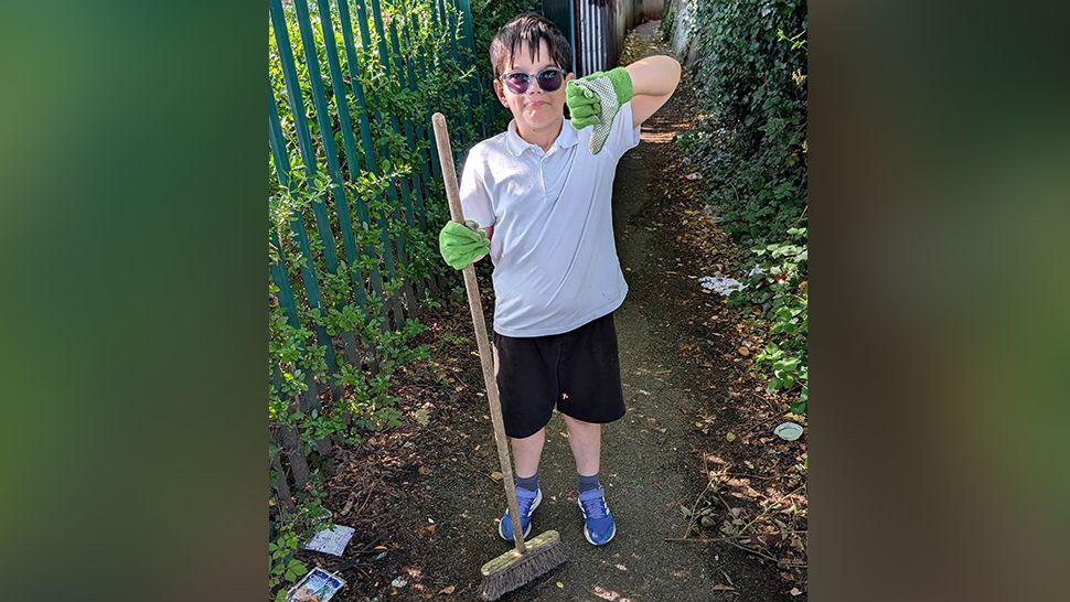 Eight-year-old Jackson wearing sunglasses, a white polo top, black shorts and green gardening gloves is standing in an alleyway with glass and rubbish. He is holding a brush and giving the thumbs down. There is a metal fence behind him with overgrowing leaves on both sides of the path. 