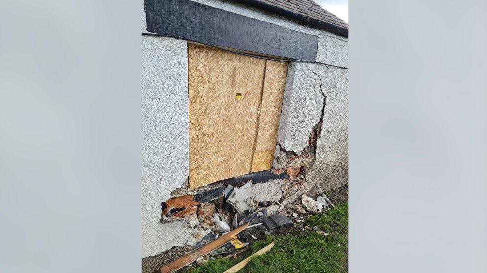 Part of the outside wall below the boarded window of the inn is smashed with its bricks exposed. There is rubble on the grass.