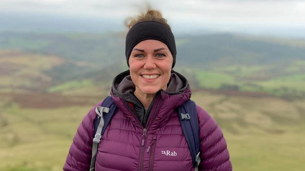 A headshot of a lady standing on a hill, she is smiling into the camera and wearing a purple puffer coat. She has a black headband on and her hair is tied up into a bun on the top of her head. She is wearing a backpack. 