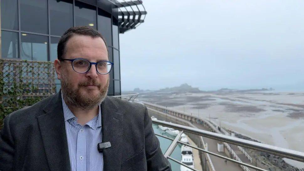 Chris Parker is standing on a balcony outside a dark glass-walled building. He is wearing a striped shirt with a jacket. He has dark short hair, a beard and a moustache and is wearing glasses. Below and behind him is an inlet with a few boats moored on it and the beach stretching to the sea beyond. It is foggy and grey.