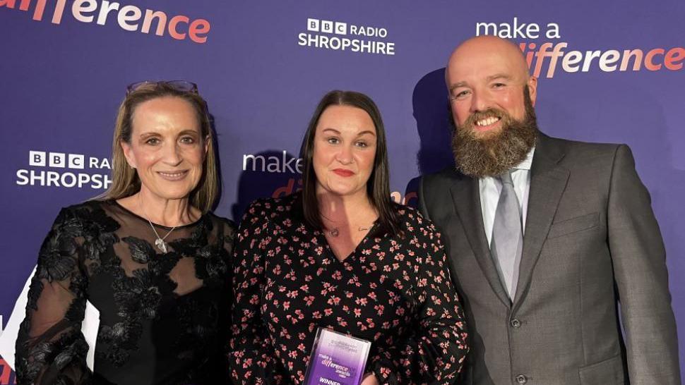 Two women and one man in front of a purple background, with one of the women holding a small purple award