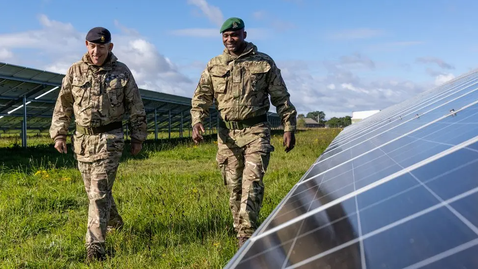 Two members of the British Army, dressed in their camouflage uniforms, walking on grass past a line of solar panels. 