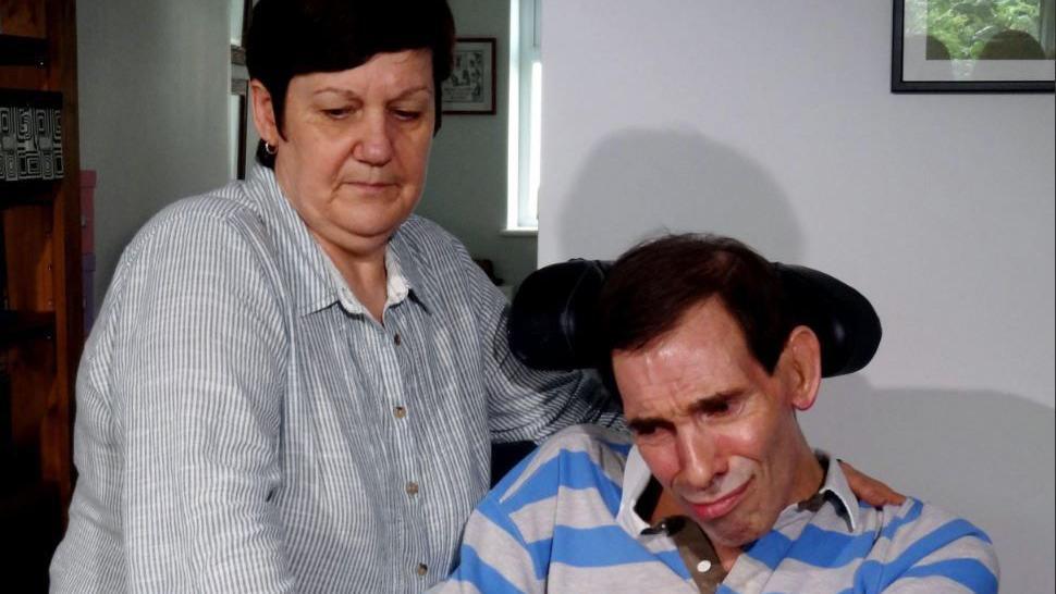 Jane stands next to her husband, who is using a wheelchair and is wearing stripy shirt. 