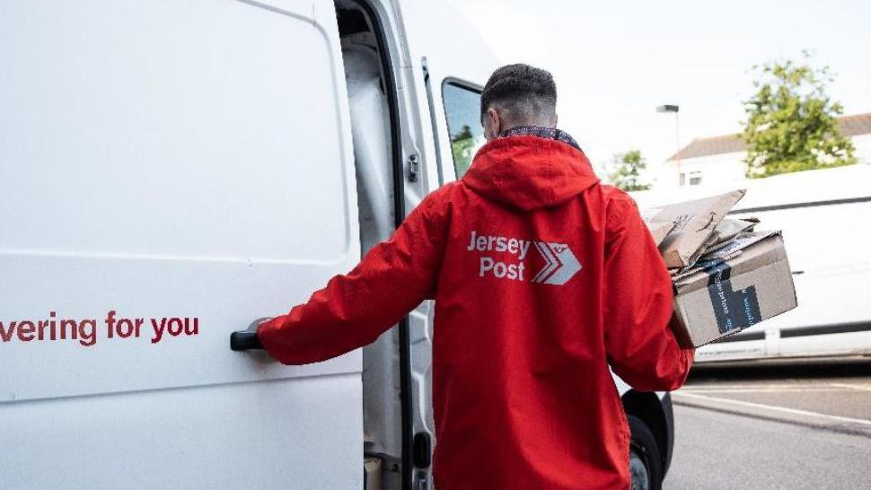 A Jersey Post postman is seen from behind, sliding closed the door to his white postal van. He is wearing a bright red coat with the Jersey Post logo on the back. He is carrying a stack of parcels. 