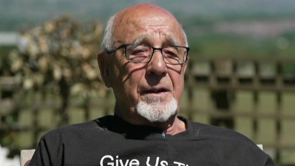 Head and shoulders image of John Morris sitting in the garden in the sunshine in a black tshirt. He is wearing glasses and has short white receeding hair and a goatee