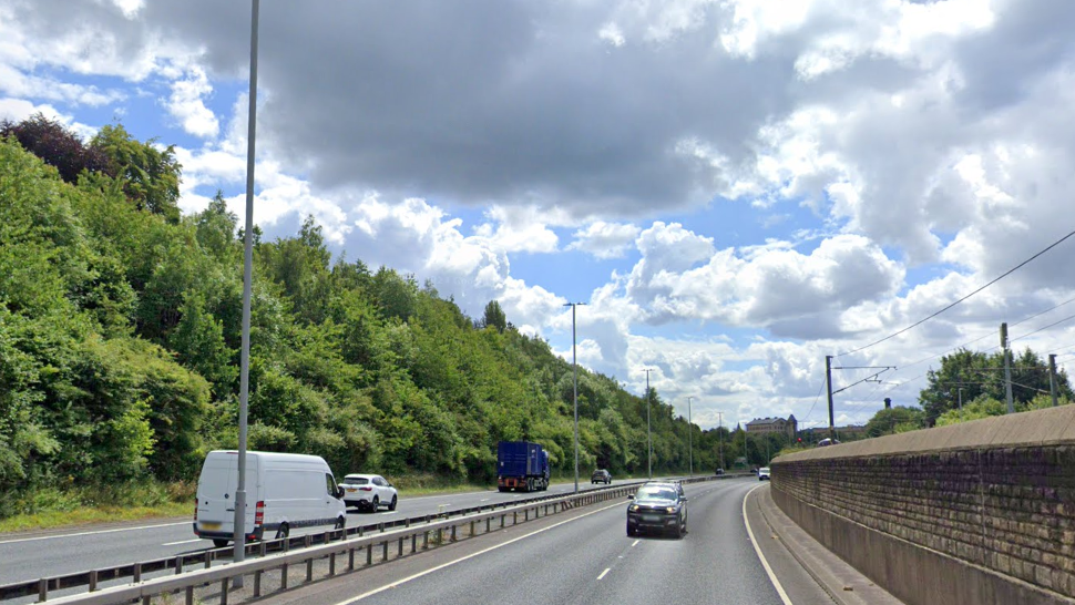 A white van, three cars and a blue truck drive along the A650. There is a railway line to the right and a line of trees and bushes to the left.