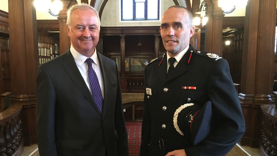 Two men, one in a dark suit and tie, the other in black formal firefighter uniform stand looking at the camera. The man in the firefighter uniform holds a formal cap under his arm.