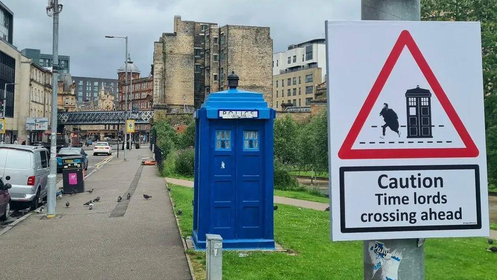 A photo of an old-fashioned blue police box. It looks like a telephone box but was used by the police in years gone by. The sign has a drawing of the Doctor Who tardis, which is a police box, and the doctor nearby. It says Caution: Time Lords crossing ahead.