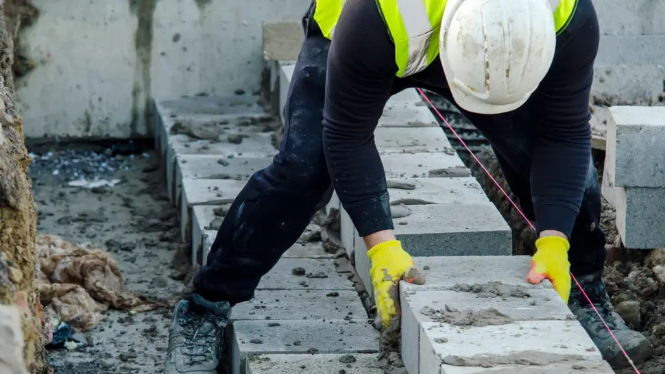 A builder places a breeze block in position in the construction of a wall on a housing scheme - he is wearing a high-viz jacket and helmet