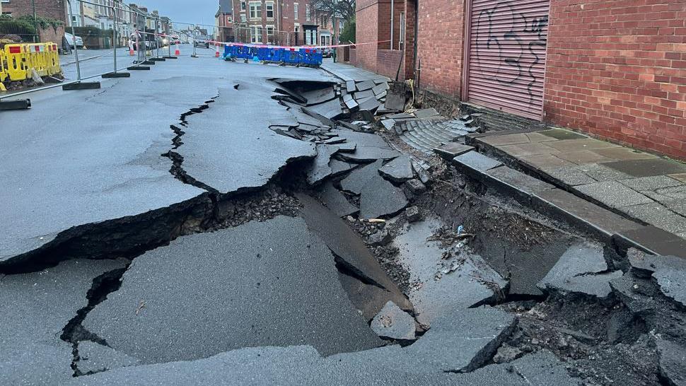 Grey temporary fencing and fluorescent barriers surround the sinkhole. The grey tarmac road is splintered and sunken.