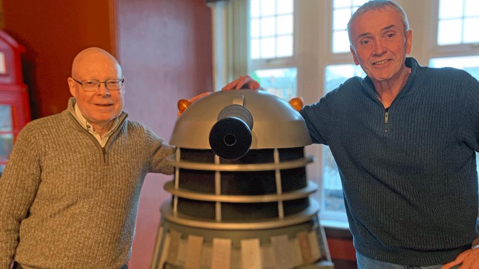 Two men in zip top jumpers - one bald headed with glasses and the other with grey hair - stand either side of a model of a Dalek from TV's Dr Who show