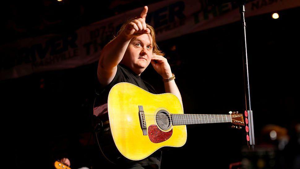 Capaldi, who is wearing a black t-shirt, is on a stage and has an arm raised and with his hand is pointing to something in the distance. He has a yellow acoustic guitar slung by a strap over his shoulders. There is a microphone stand next to him.
