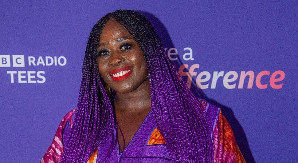 Susan Mansaray has black and purple braised hair. She's wearing red lipstick and has a bright pink, purple and orange top on.