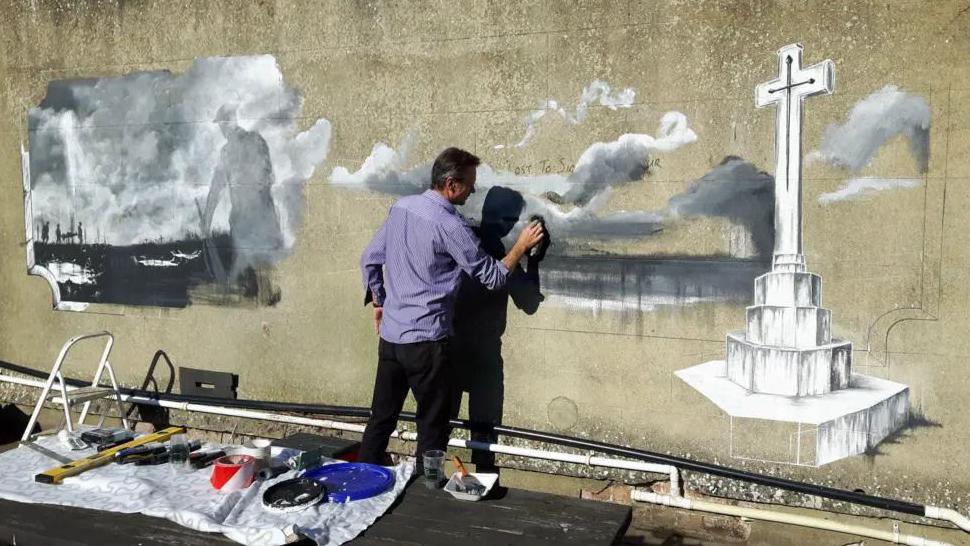 A man with hs back to camera painting a mural on a wall with his paints behind him