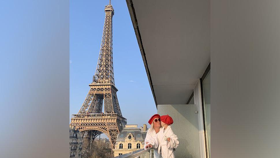 Hayley and Jamie wearing red berets standing on a balcony in an apartment block with the Eiffel Tower in Paris just behind them