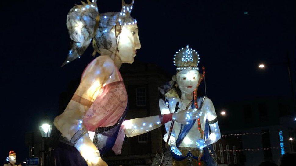 Large lighted puppets, one representing a man with a crown and other a woman with her hair tied.  They are towering above a crowd in a parade.