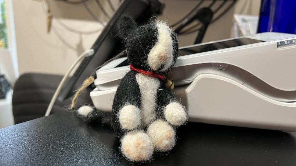 A small soft toy resembling a black and white cat on a black desk next to some office equipment.