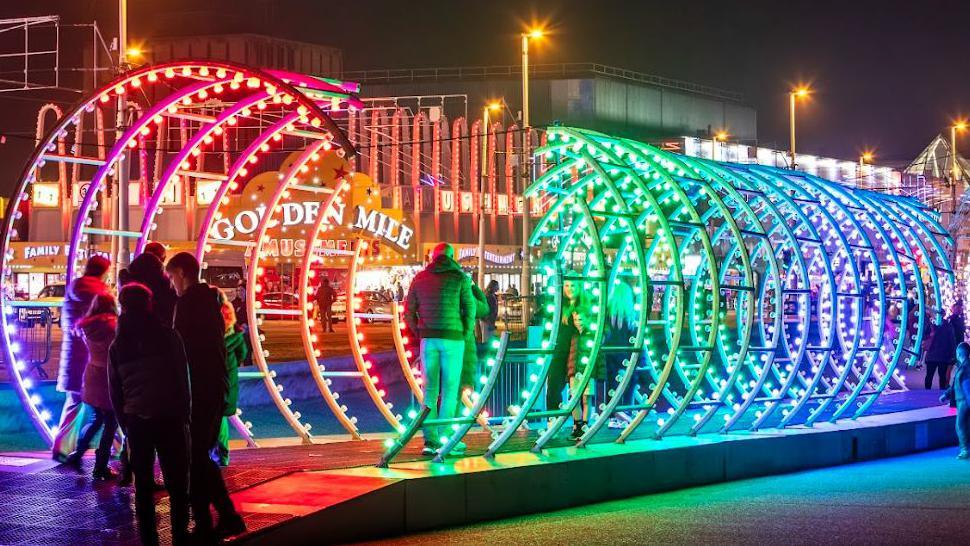 A large light installation, in rainbow shades, resembling a giant Slinky. People are standing at one end and a man is walking through it.