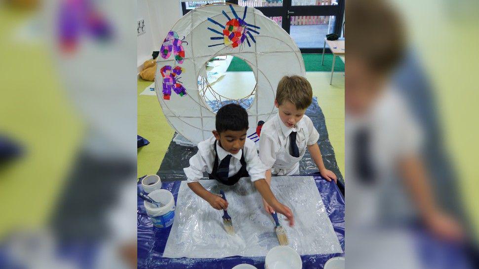 Two children, each with a brush, pasting glue onto a white square of material with a large lantern behind them, decorated with butterflies and bugs