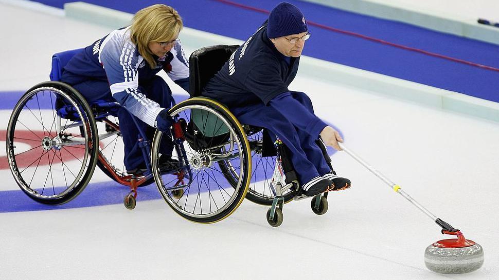 Angie Malone z Velké Británie drží invalidní vozík Michaela McCreadieho, když doručuje kámen v curlingu na led pomocí tyče.