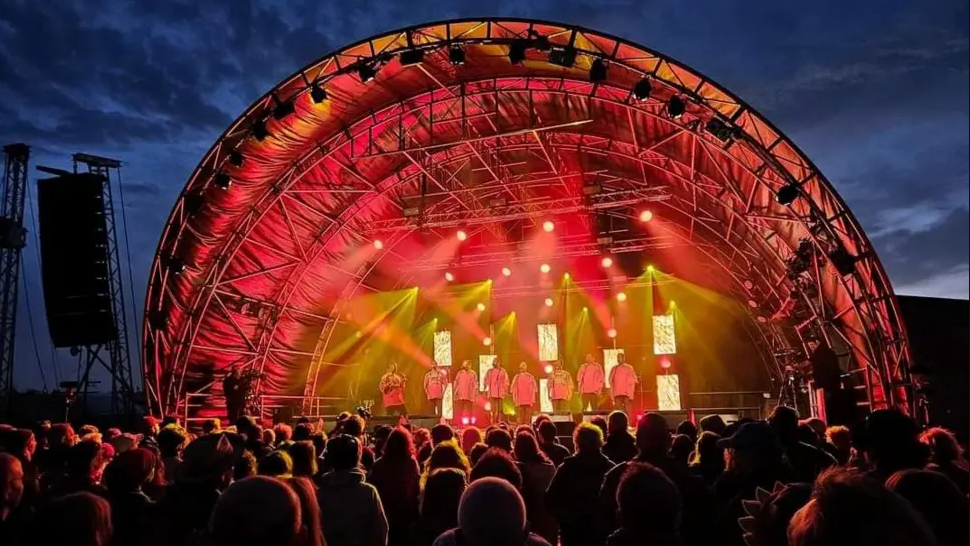 A dome-like structure stands over the stage with red and yellow lights. The night sky can be seen behind it with a crowd of people stood in front of the stage.