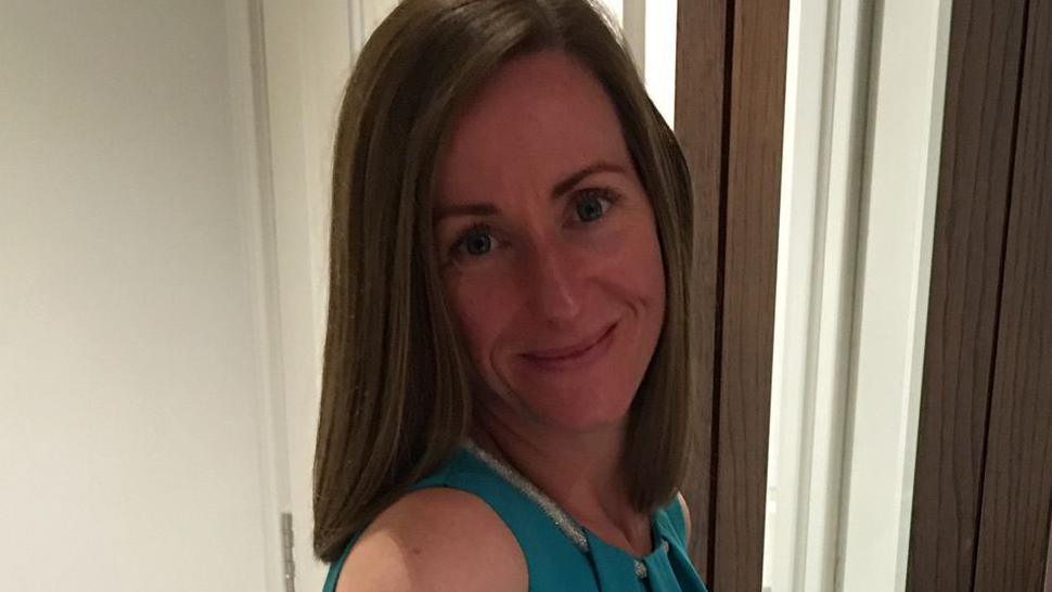 A woman in a blue dress is smiling, posing for the camera. She has brown hair and is stood next to a mirror.