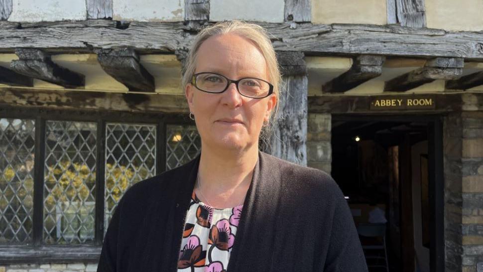 A woman with blonde hair, glasses, a flowery top and a grey cardigan stood outside a building with a lead-framed window