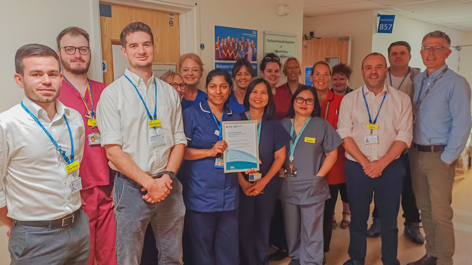 A group of male and female doctors and nurses, some in smart casual clothes and some in hospital scrubs, line up for the camera at the Royal United Hospital in Bath. In the centre two nurses in blue scrubs are holding up an award in a frame